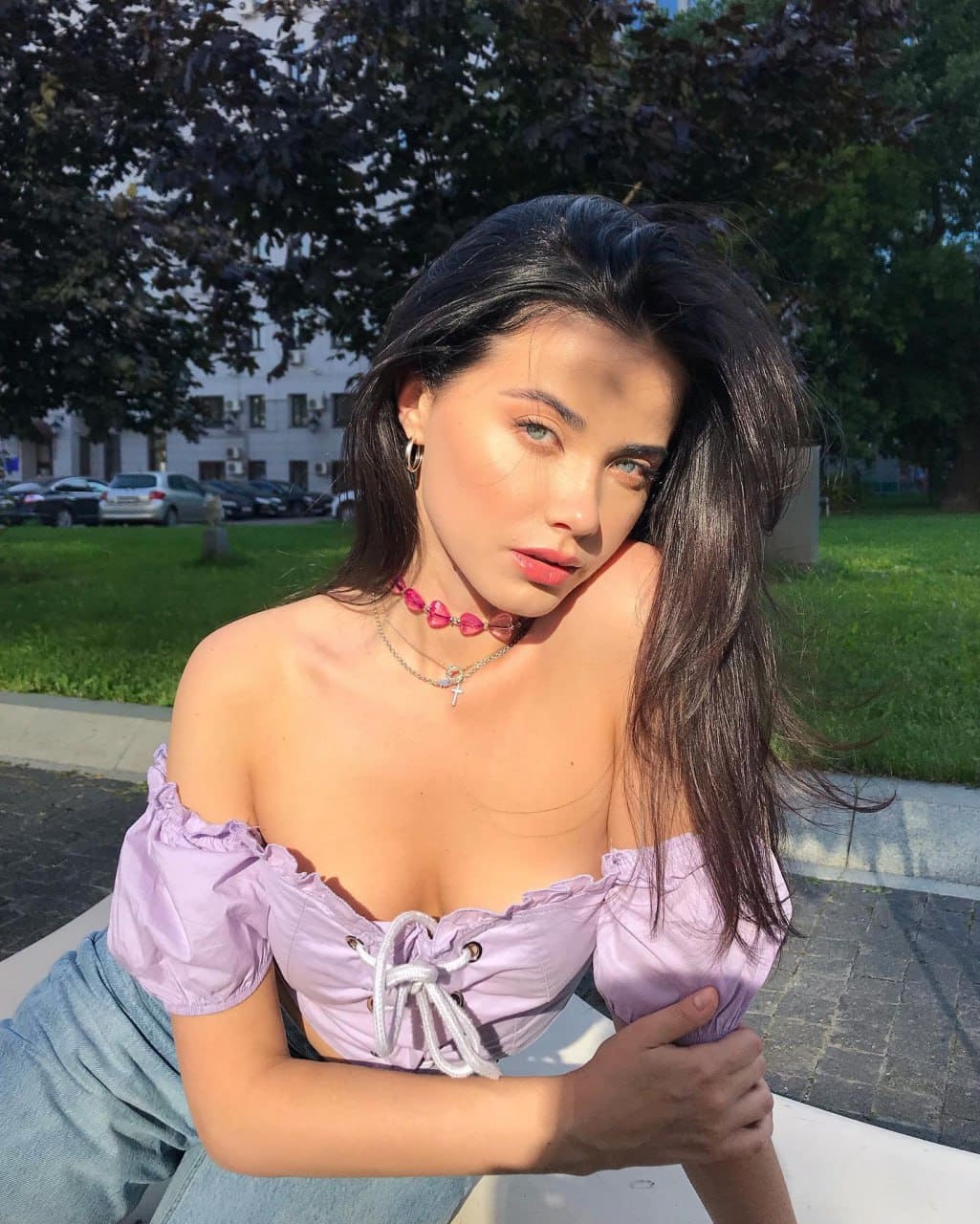 A woman in a light purple off-shoulder top and blue jeans sits on pavement outdoors, wearing a pink choker against a background of grass and trees.