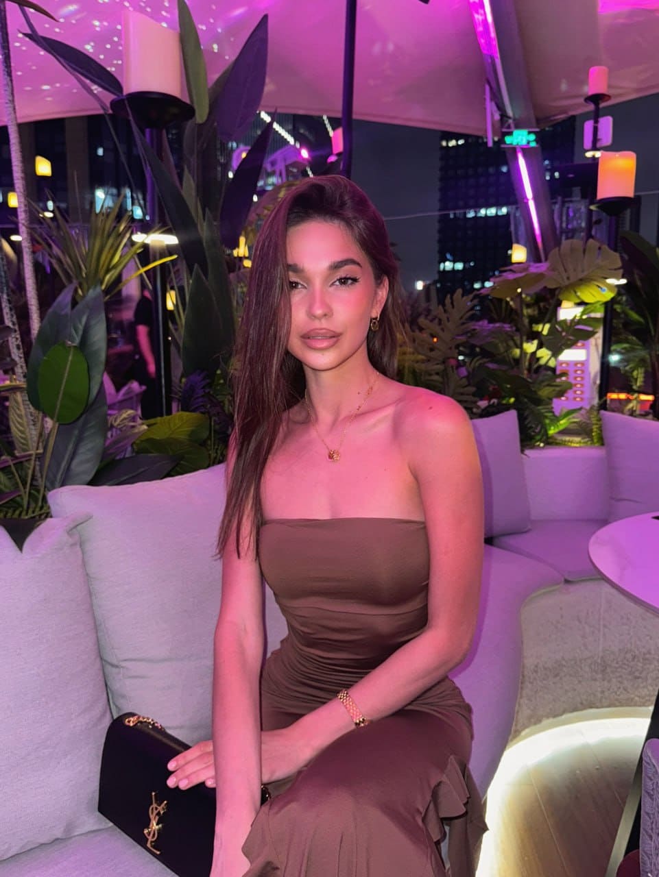 A woman in a brown strapless dress sits on a couch in a plant-filled lounge, holding a black clutch on her lap.