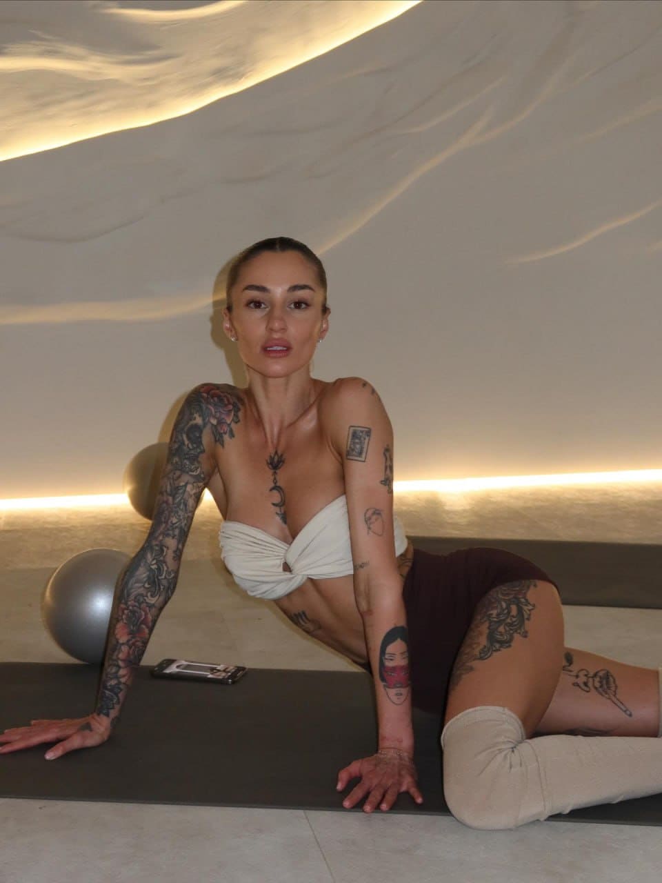 A tattooed woman sits on a black exercise mat in a white strapless top and dark shorts, with a silver exercise ball and phone beside her.