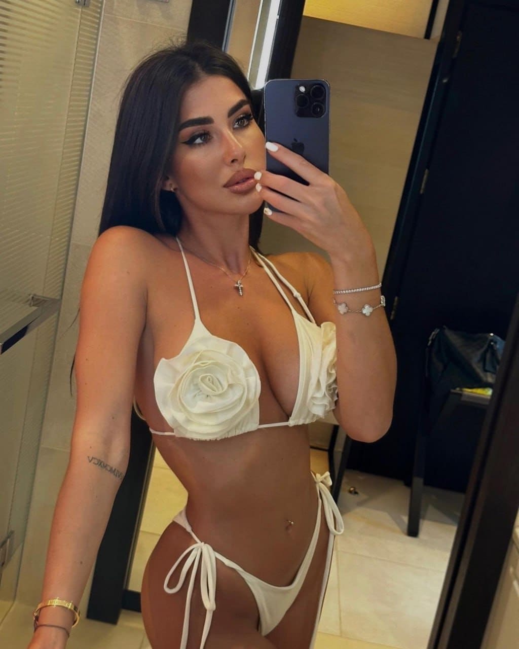 A woman takes a mirror selfie in a white floral bikini, holding a dark smartphone in a tiled room with a glass door.