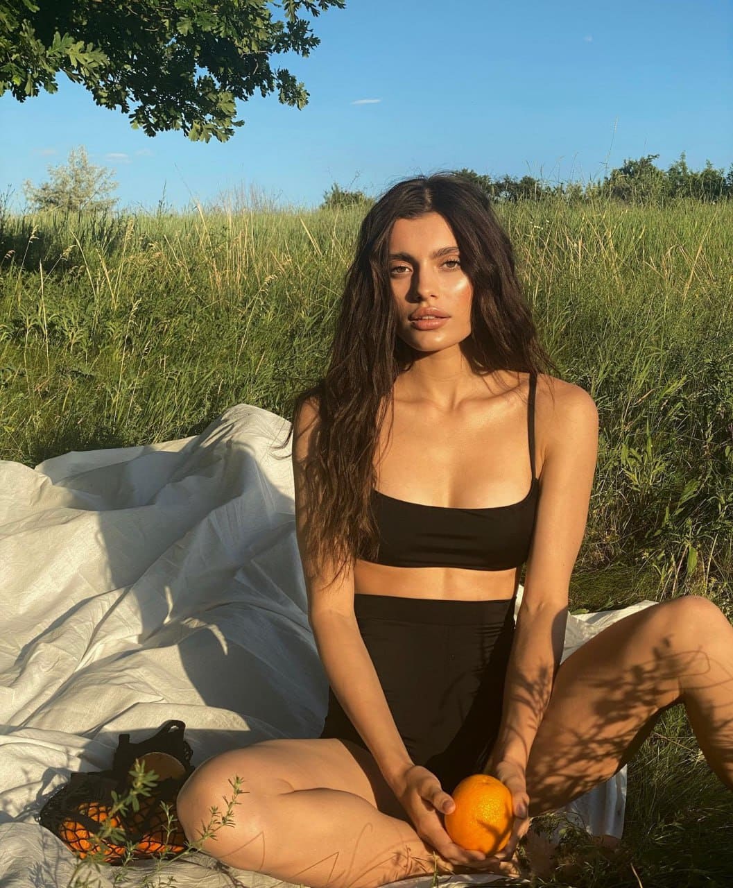 Dominica sits cross-legged on a white sheet in a grassy field, wearing a black sports bra and shorts while holding an orange.