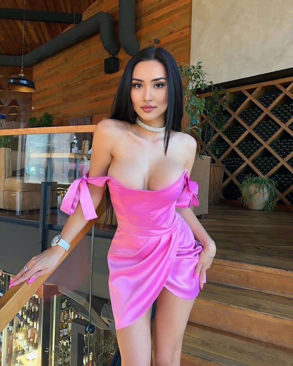 Lisanne in a pink satin off-shoulder dress with bow details, pearl choker, and watch, leaning on a wooden railing in a wood-paneled room.