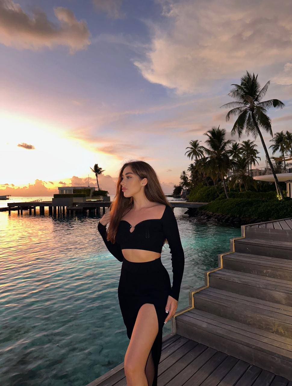 In profile on a wooden deck by a pool, a woman wears a black long-sleeved two-piece outfit against a sunset backdrop with palm trees.