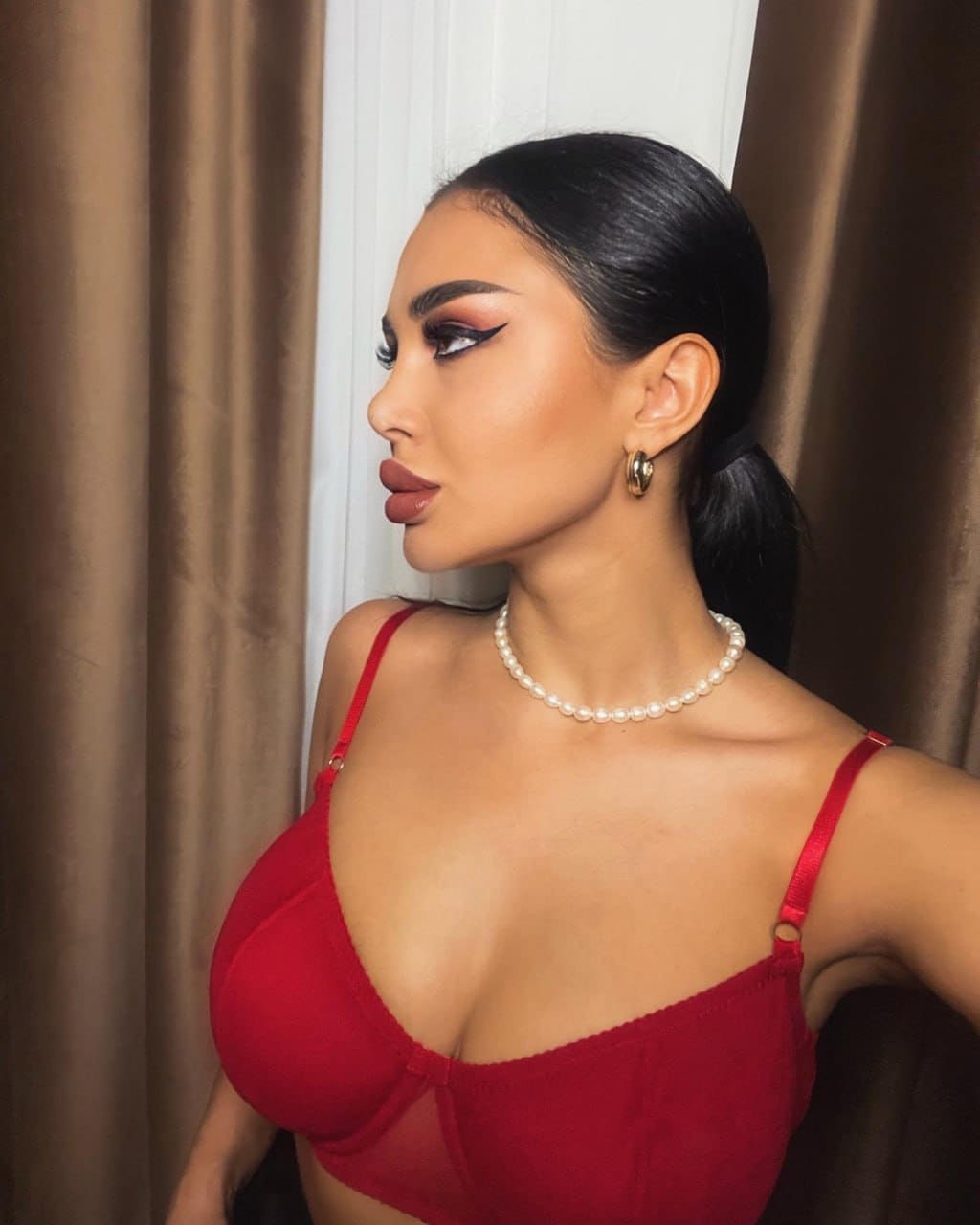 A woman with a ponytail stands in profile against brown curtains, wearing a red bralette, pearl necklace, and gold hoop earrings.