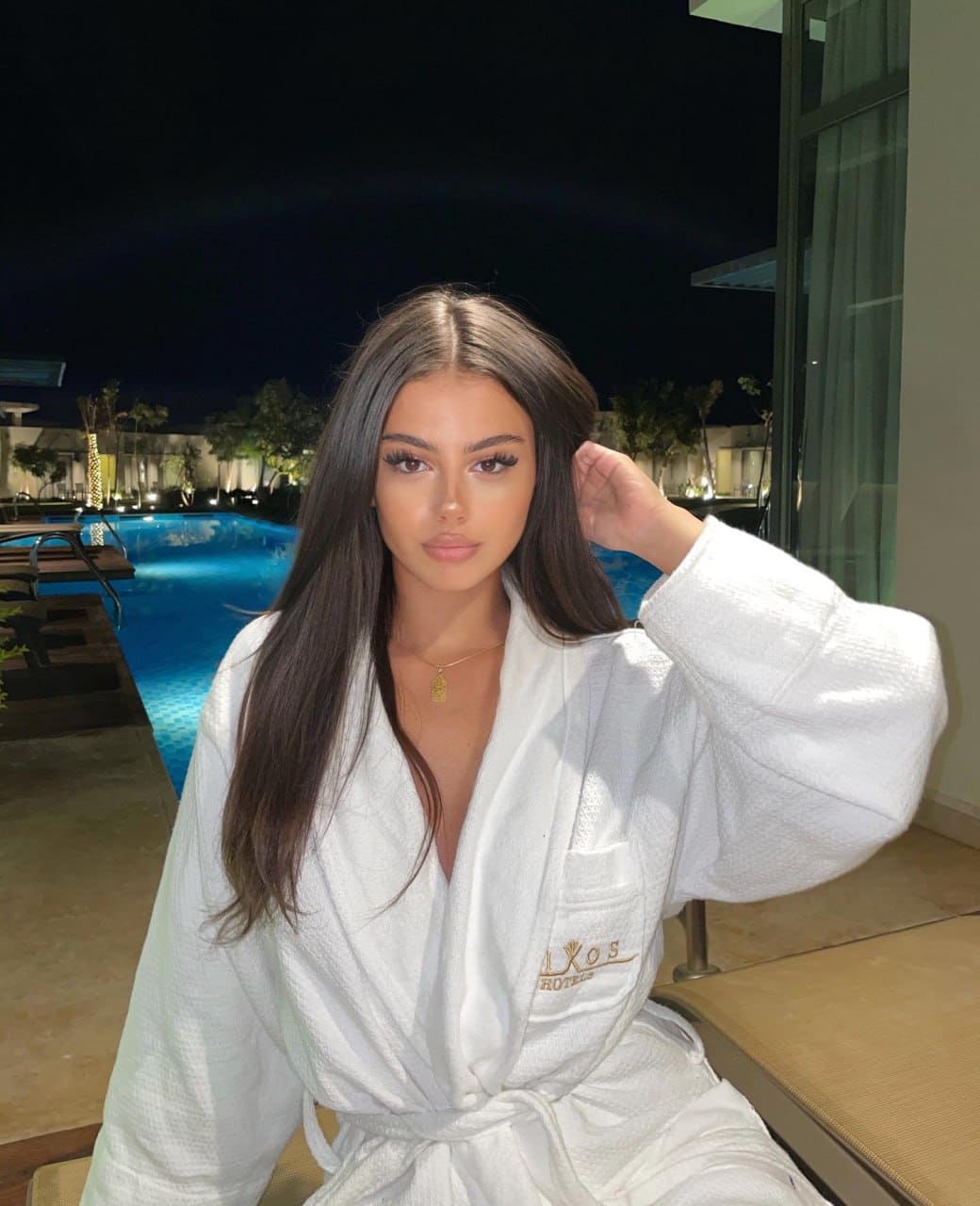 A woman with long dark hair sits by a lit swimming pool at night, wearing a white waffle-knit bathrobe tied at the waist, hand raised to hair.