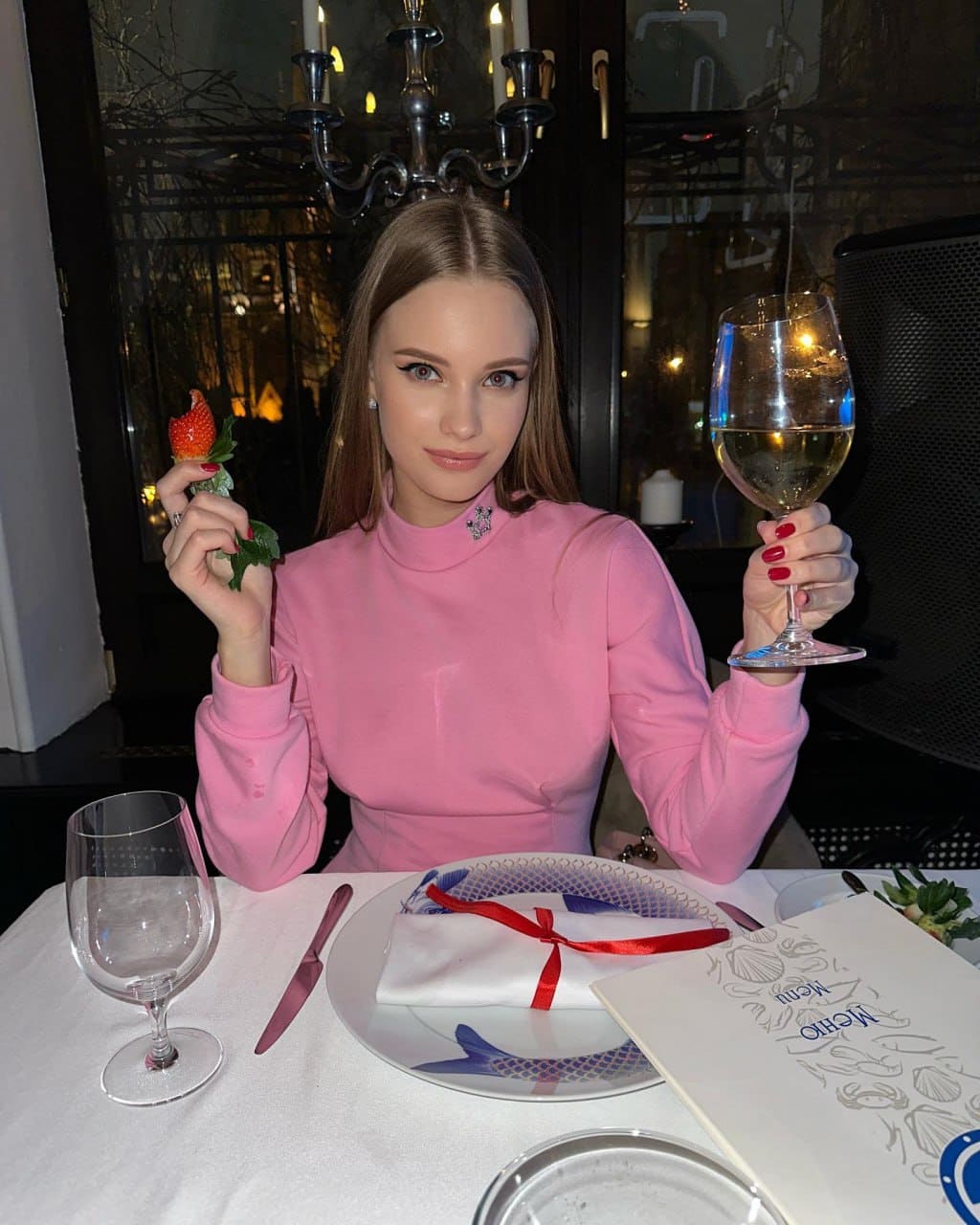 Milena sits at a restaurant table in a pink turtleneck, holding a wine glass and strawberry, with a chandelier behind her.