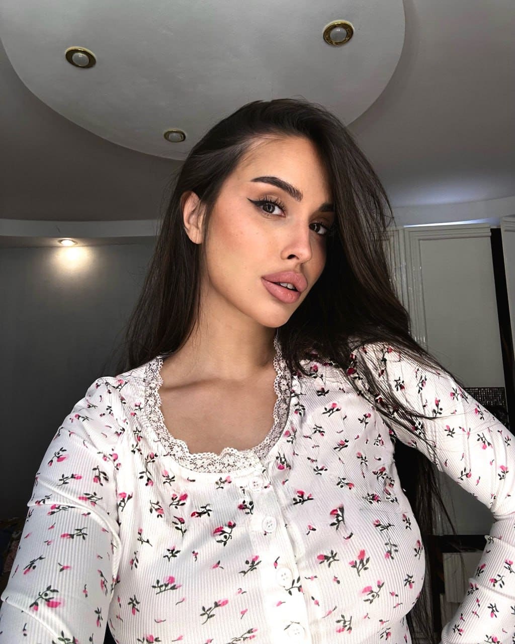 A woman in a white floral blouse with lace trim poses indoors under a recessed ceiling light.
