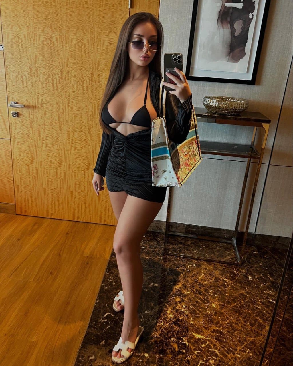 In a mirror selfie, she wears a black cutout dress and holds a phone and tote bag on a marble floor.