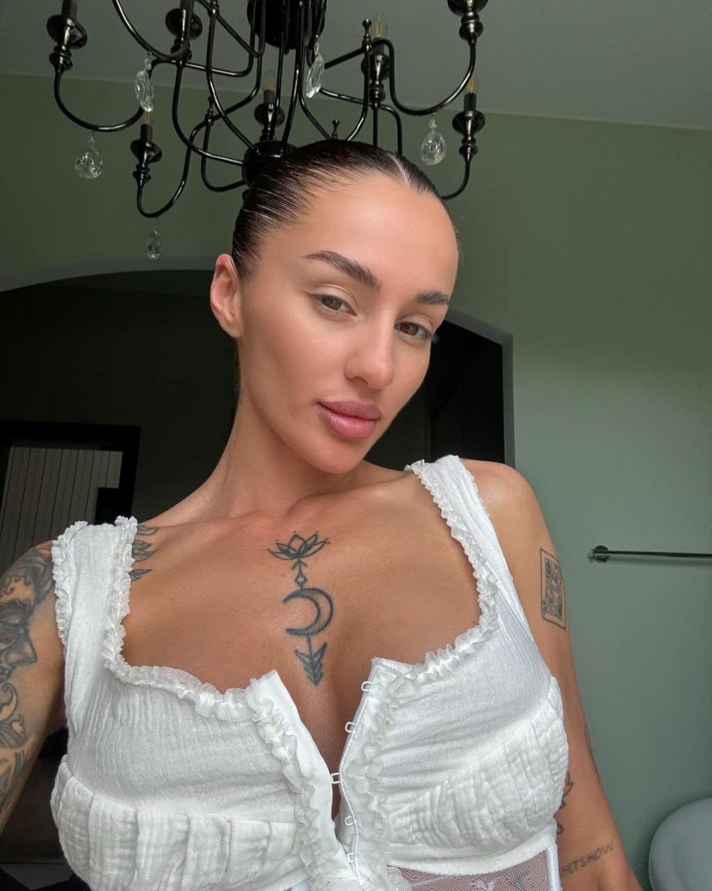 A woman with slicked-back hair poses indoors in a white ruffled top with a deep neckline, standing beneath a black chandelier with visible tattoos.
