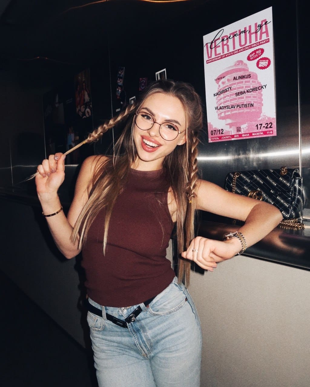 A woman with braided blonde hair and glasses smiles in a brown top and jeans, leaning on a counter near a pink poster.