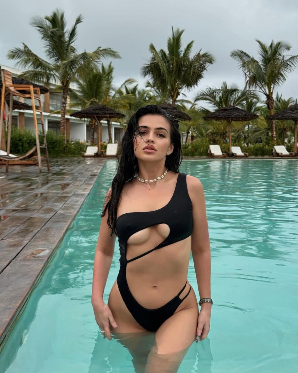A woman stands in a turquoise pool in a black cutout swimsuit, with palm trees and umbrellas behind her.