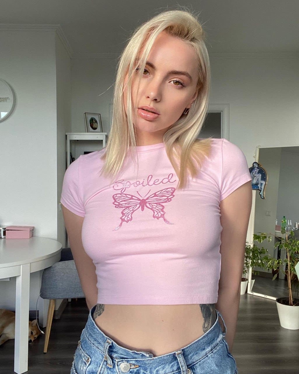 A blonde woman wears a pink butterfly crop top and low-rise blue jeans, standing indoors near a white table, chair, and potted plants.