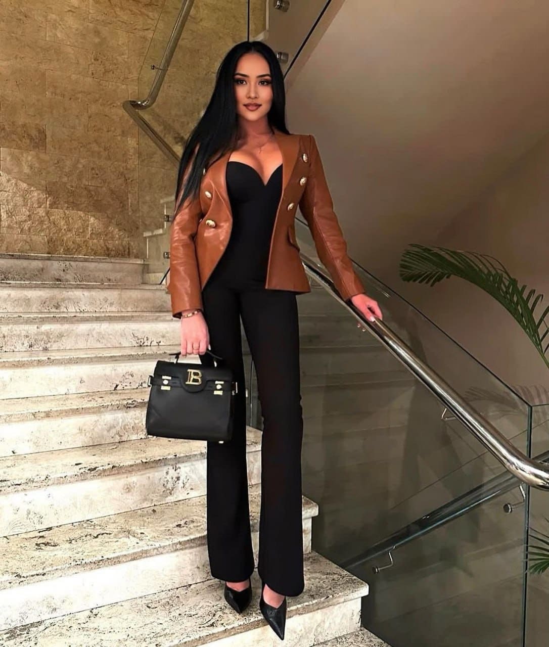 On a marble staircase wearing a brown leather jacket over a black top and trousers, holding a black handbag while resting a hand on the railing.