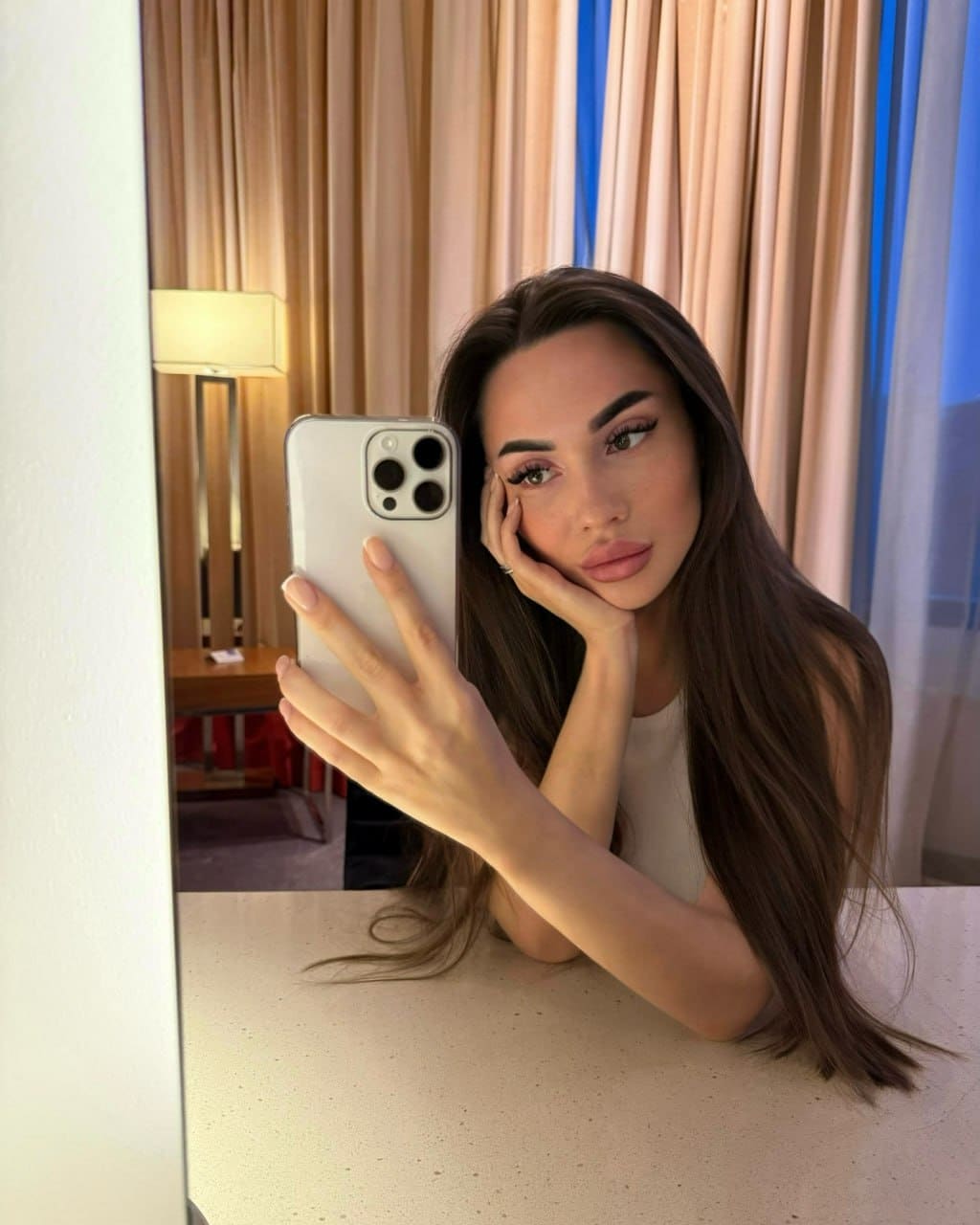 A mirror selfie shows a woman resting her chin on her hand, wearing a light sleeveless top against beige curtains.
