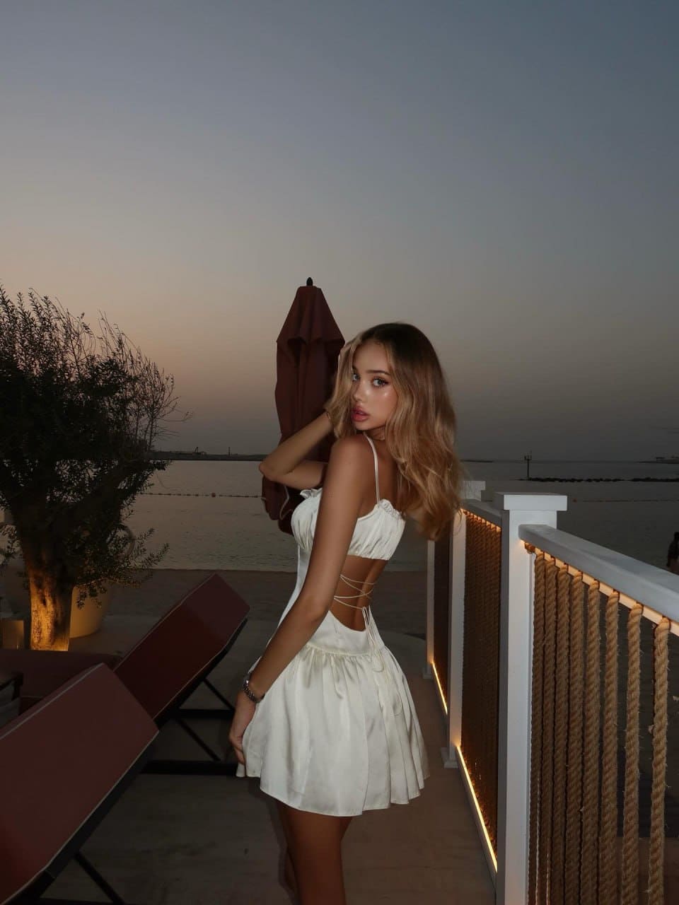 At dusk on a waterfront terrace, a woman in a white two-piece outfit rests her hand near her neck beside a rope balustrade.