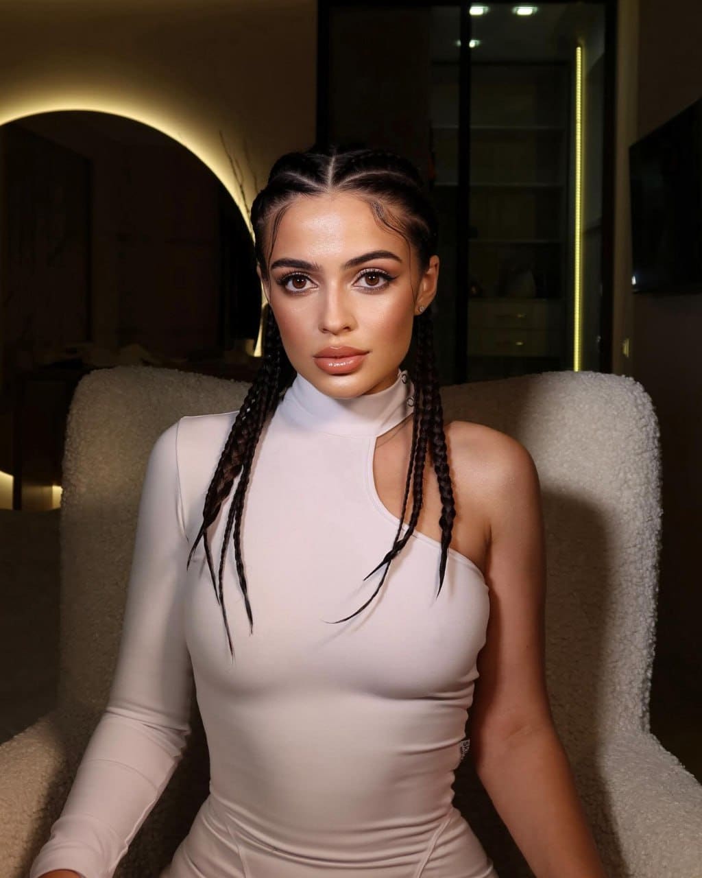 Sonia sits in a beige armchair wearing a white one-sleeve top, with long braids, in a dimly lit room.