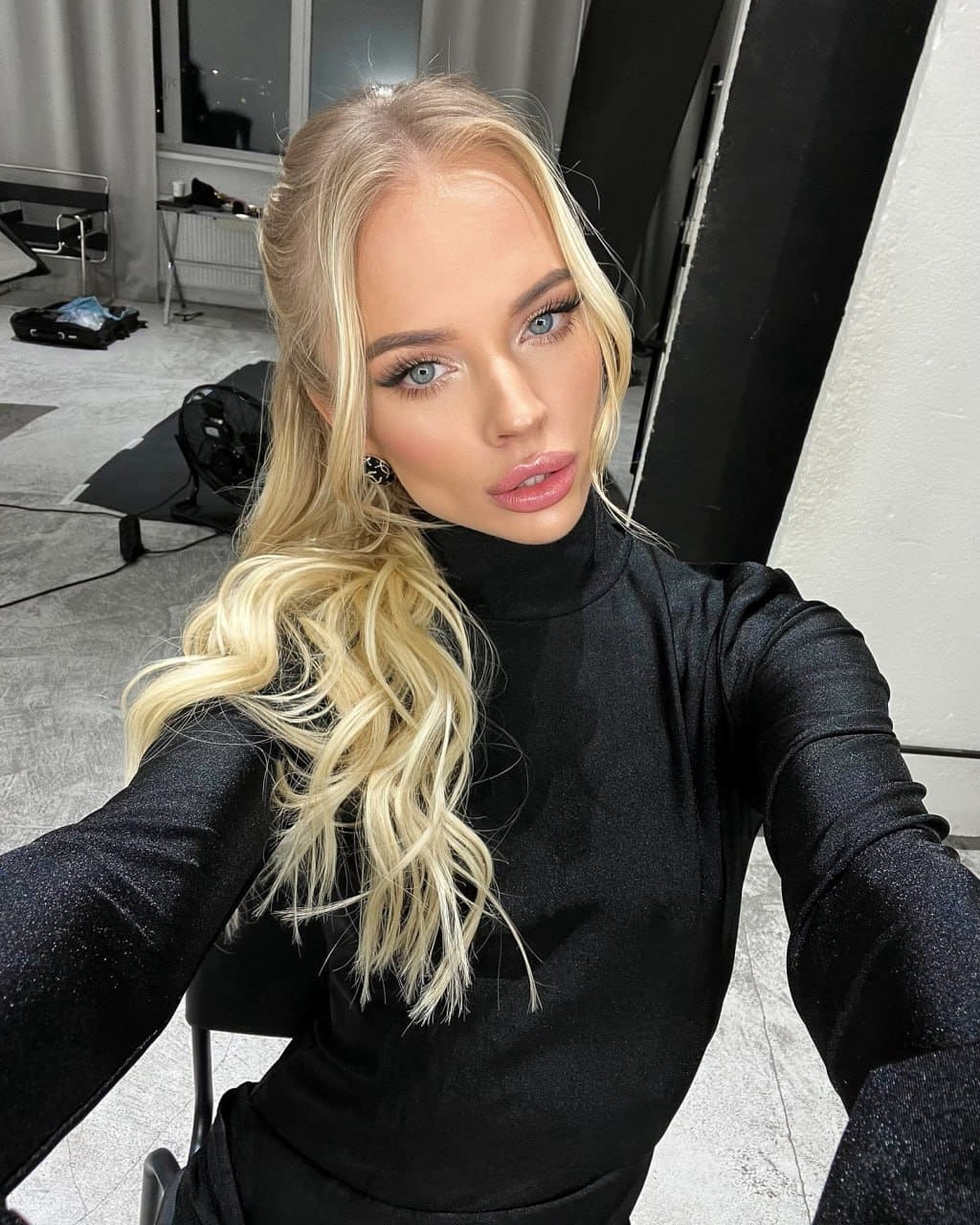 Margot takes a selfie in a studio, wearing a black long-sleeved turtleneck, with a chair and equipment visible in the background.