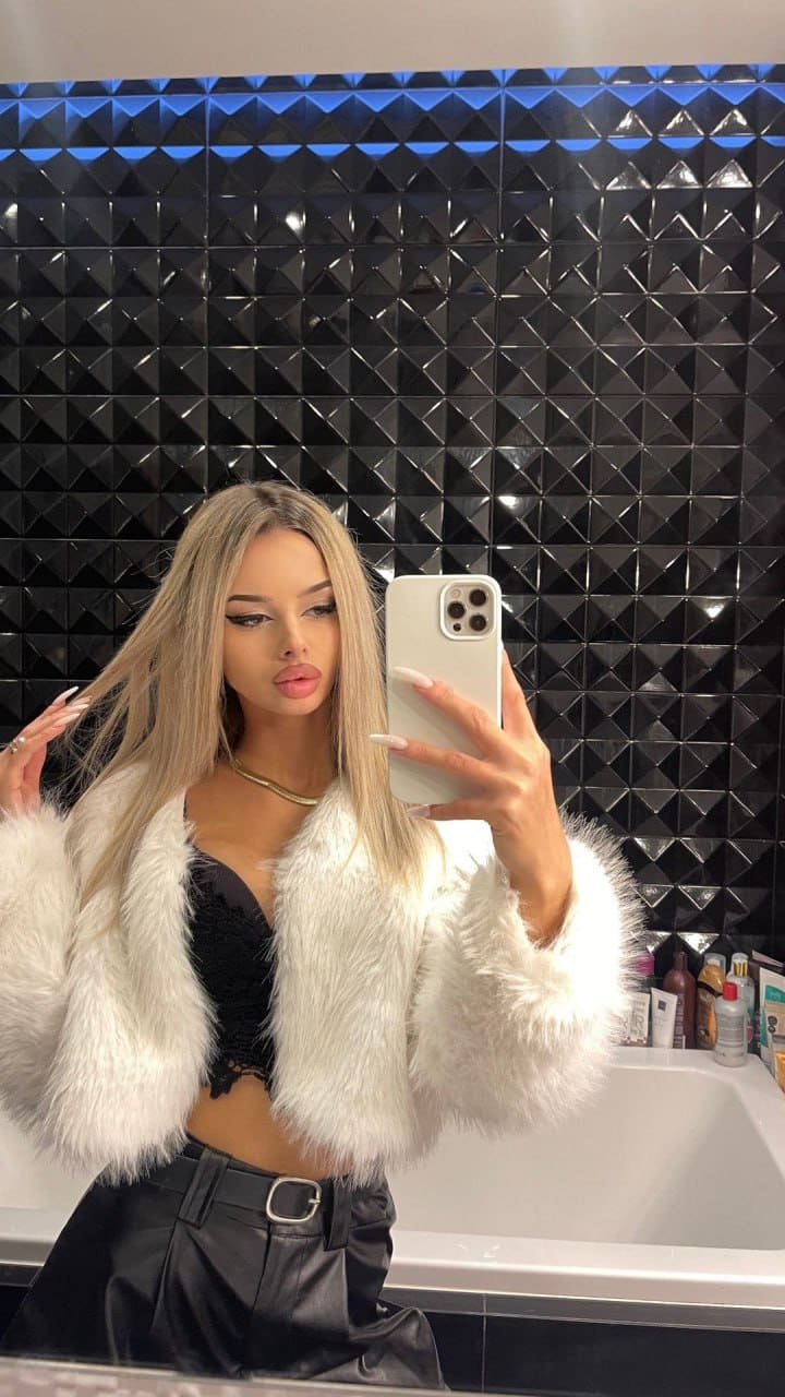 Mirror selfie of a woman in a white fluffy jacket and black leather pants against a geometric tile wall.