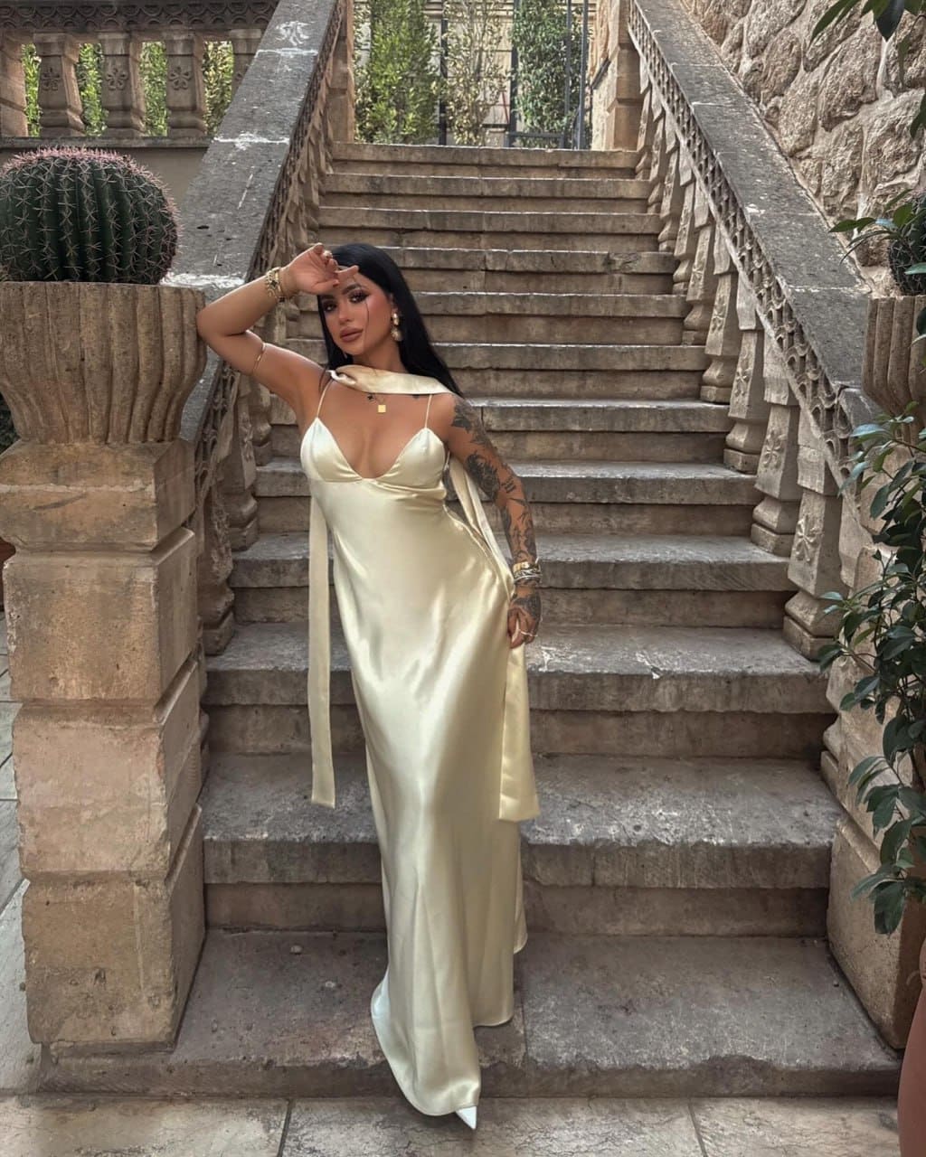 Standing on a stone staircase in a floor-length cream satin slip dress, one hand to forehead, with a potted cactus behind.