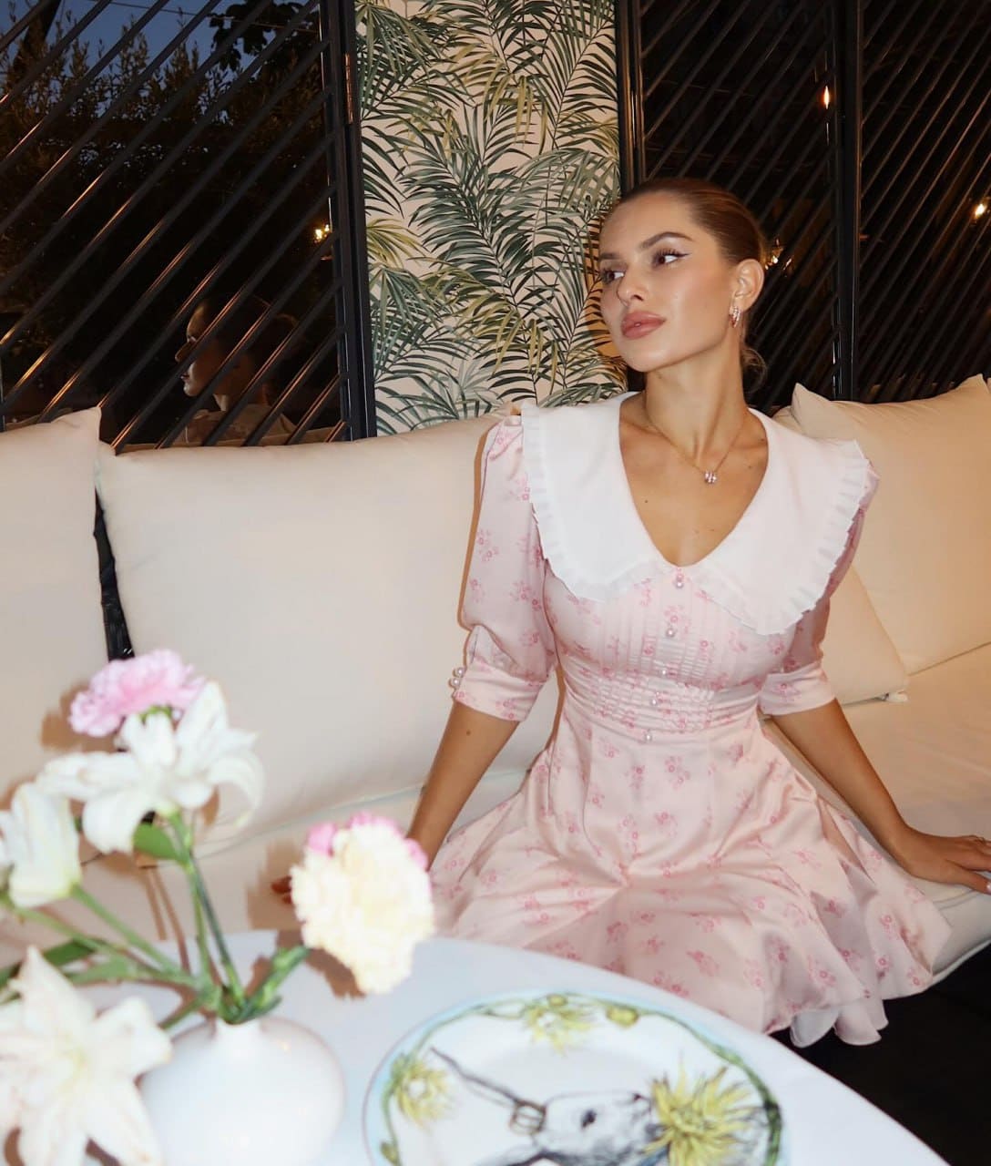 Woman in a pink floral dress with ruffled collar sits on a cream sofa, flowers in foreground, leaf-patterned wall.