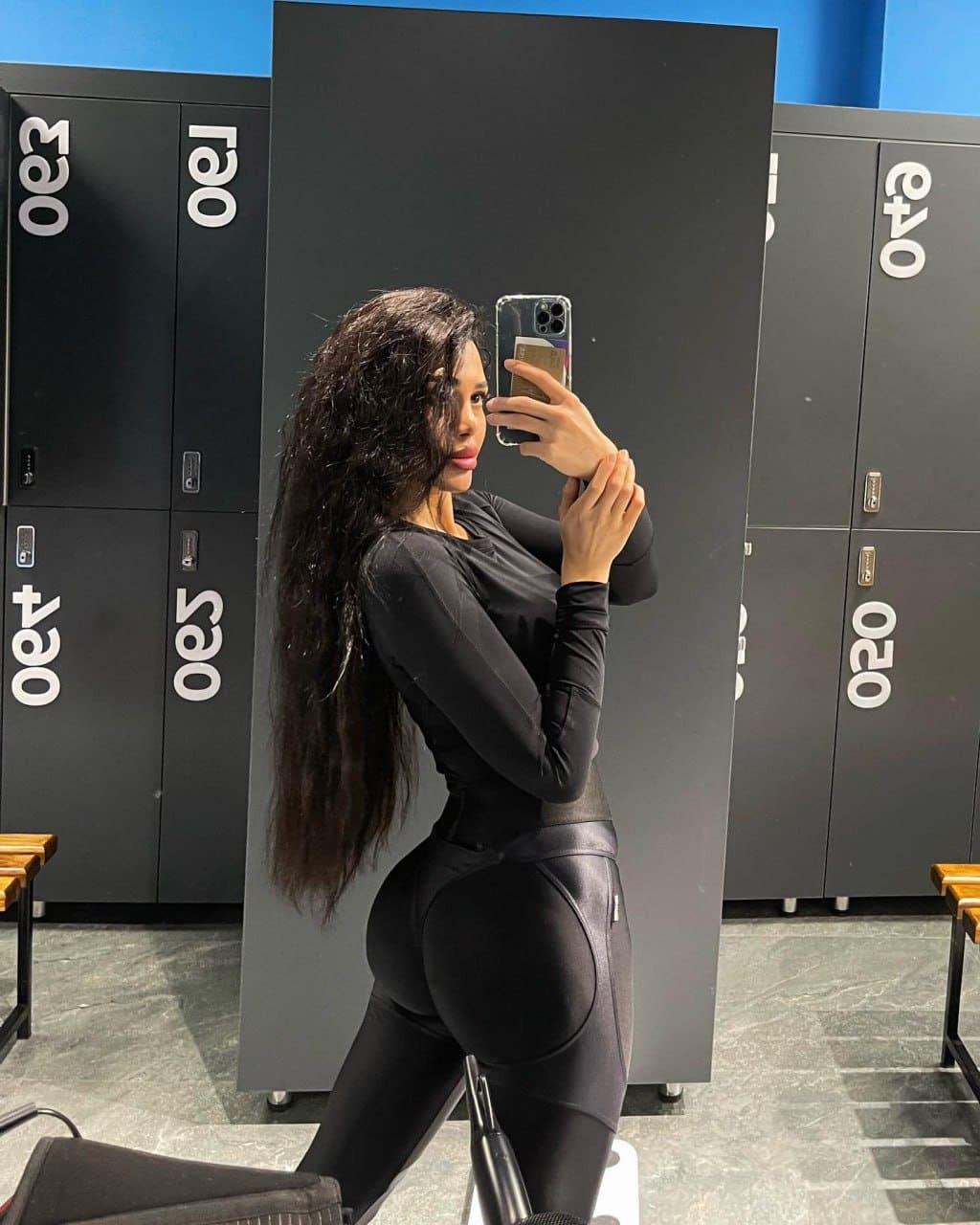 A woman in a black long-sleeve top and leggings takes a mirror selfie showing her back, standing before black lockers and a blue wall.