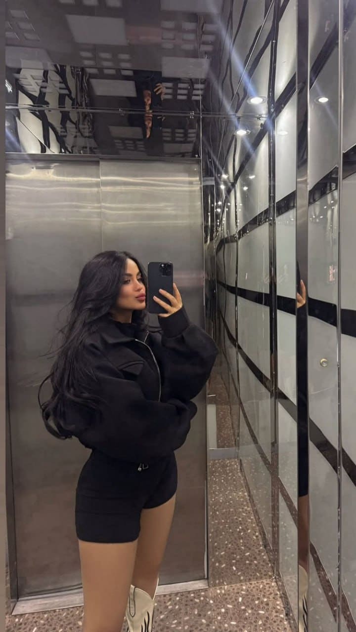 Mirror selfie in a mirrored elevator wearing a black cropped jacket, black shorts, and white boots while holding a smartphone.