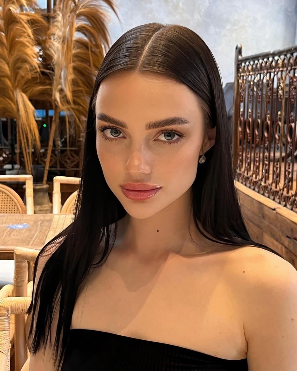 Nivia with long dark hair in a black strapless top, close-up portrait indoors with wicker chairs and dried palm fronds.
