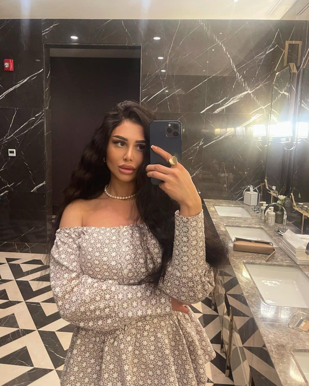 A woman in an off-the-shoulder floral dress and pearl necklace takes a mirror selfie in a marble-walled bathroom, holding a dark smartphone.