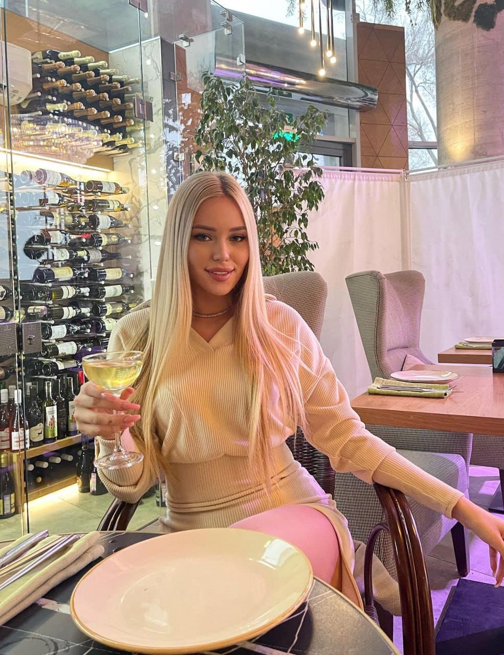 Woman in a beige ribbed top and skirt holds white wine at a restaurant table with wine cellar in background.