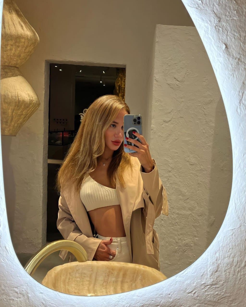 Mirror selfie in a bathroom showing a white crop top, white pants, and open beige trench coat against stone sink and textured walls.