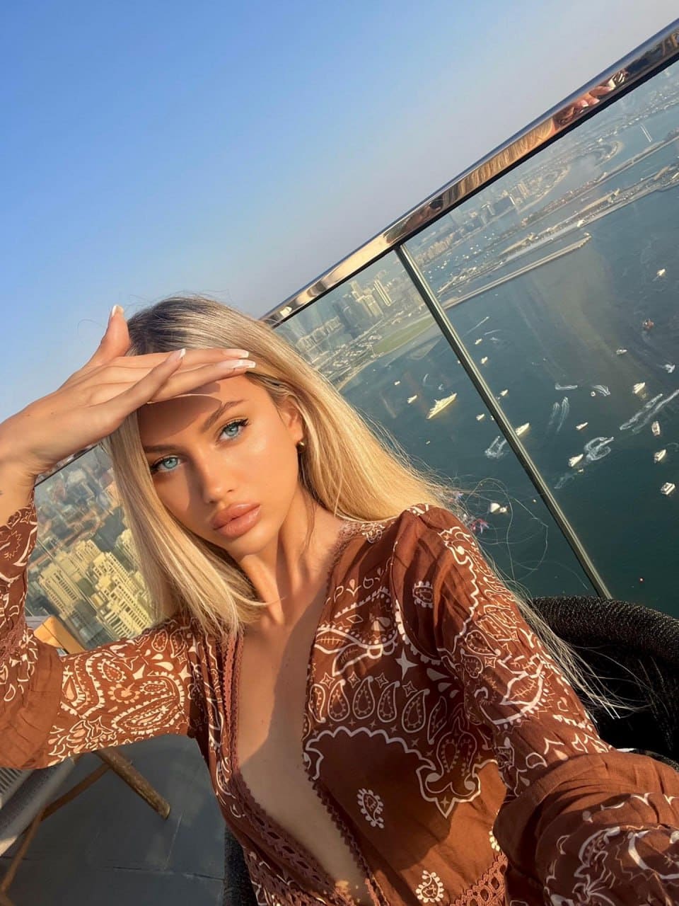 A woman in a brown paisley robe poses on a rooftop terrace, hand shading her eyes, with a city view.