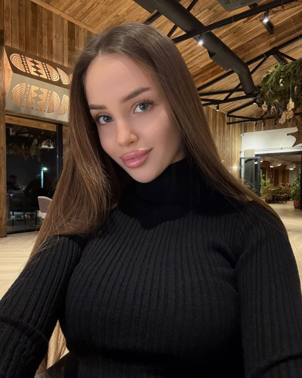 A close-up portrait of a woman in a black ribbed turtleneck sweater, set against wood paneling with wall art and hanging plants.