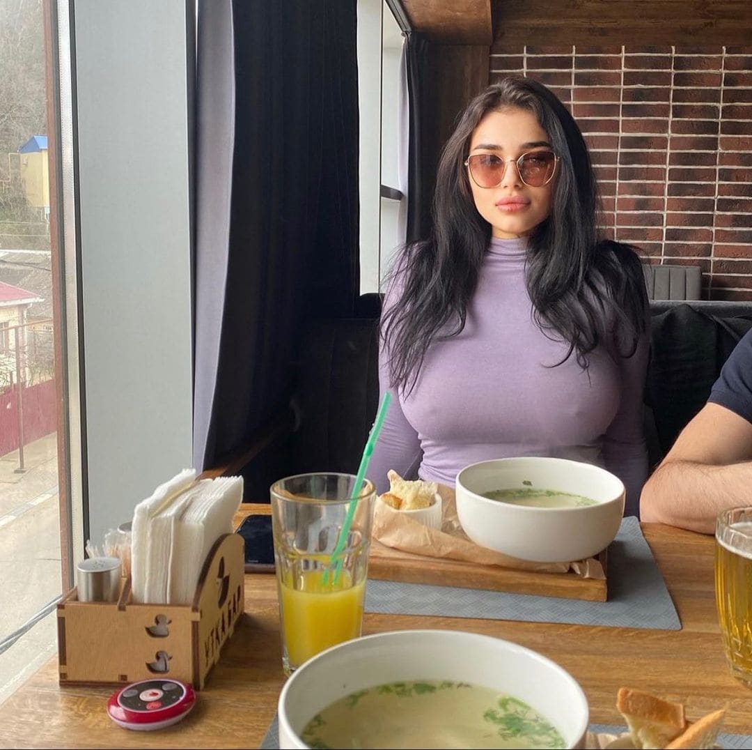 Woman in lavender turtleneck and sunglasses sits at restaurant table with soup, juice, and bread.