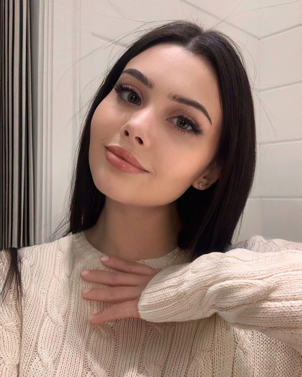 A woman in a cream cable-knit sweater rests a hand on her chest against a white paneled wall and curtain indoors.
