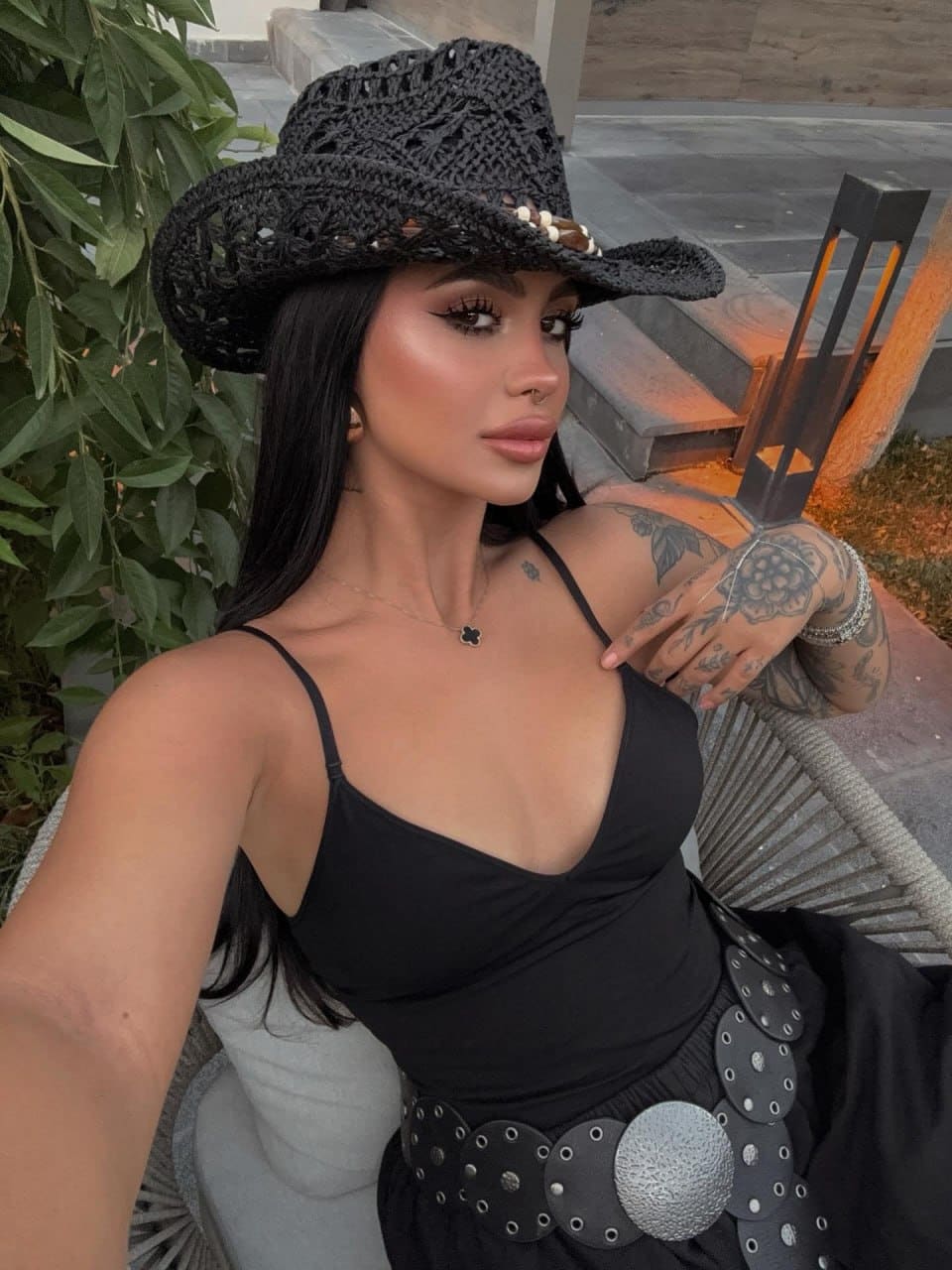 Helen wears a black crocheted hat, spaghetti-strap top, and studded belt, posing outdoors near green foliage and a stone wall.