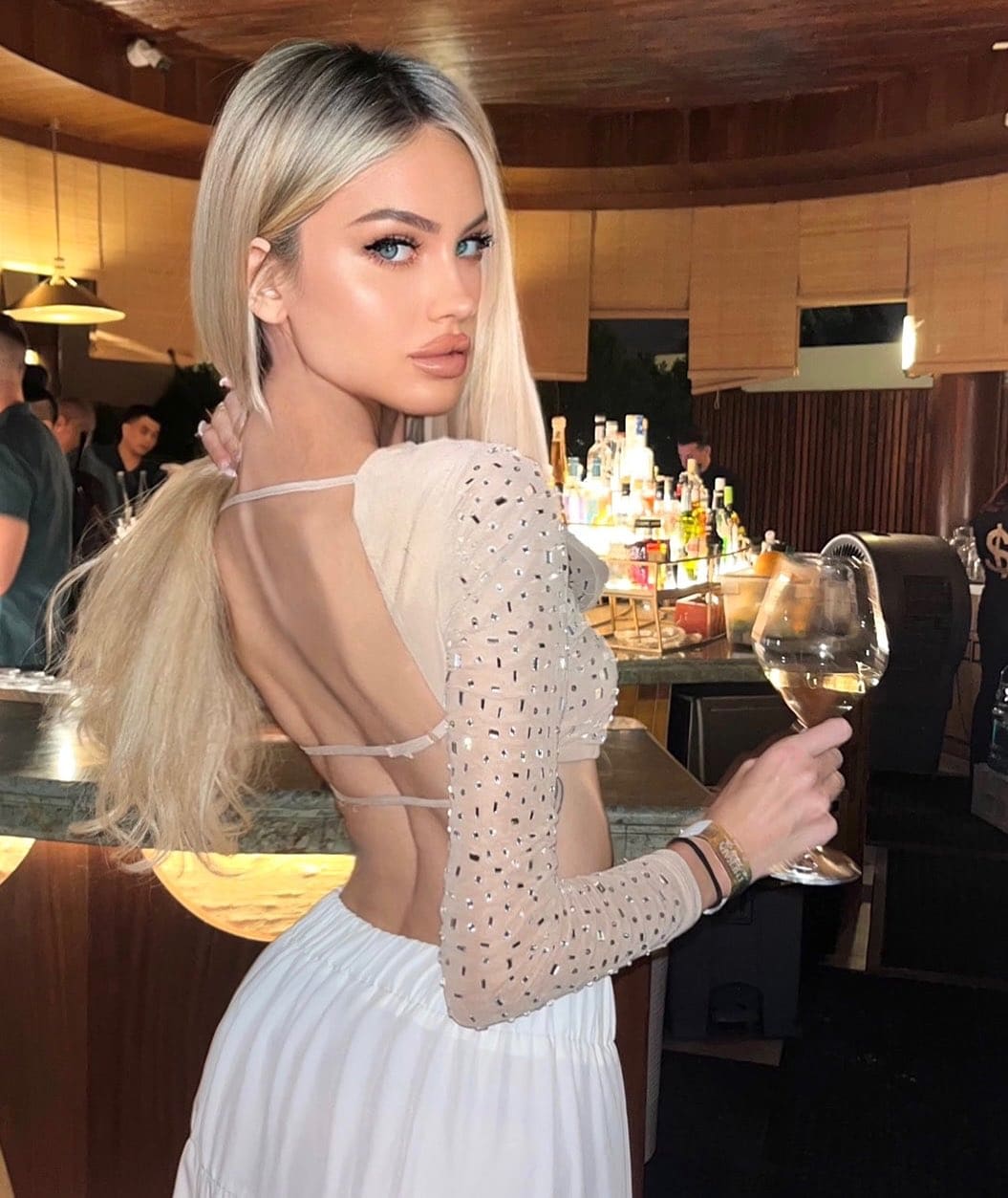 A woman with a ponytail stands at a bar in a white open-back top and skirt, holding a glass of white wine.