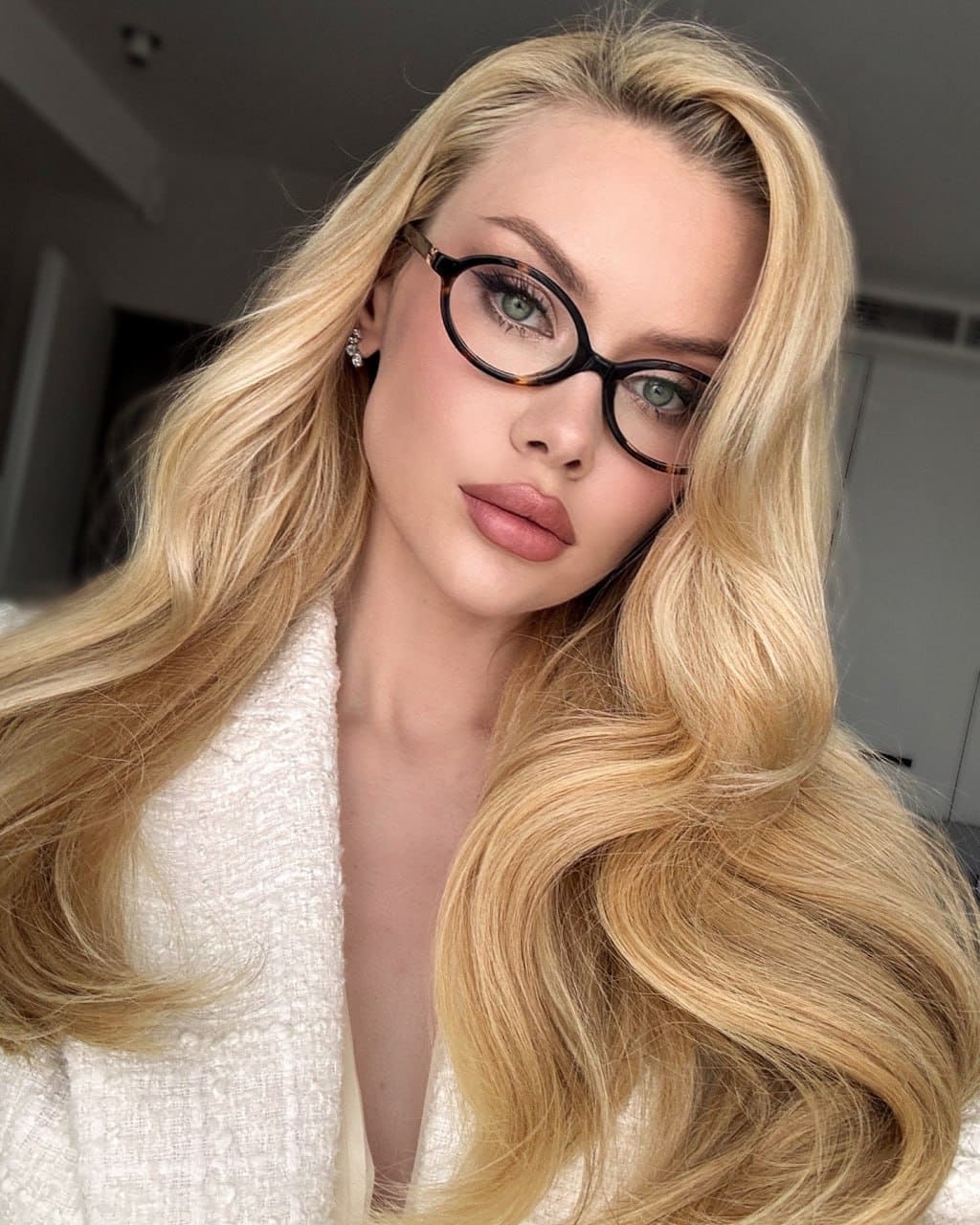 A woman with long wavy blonde hair and round tortoiseshell glasses wears a white textured V-neck top, posing indoors against a blurred background.