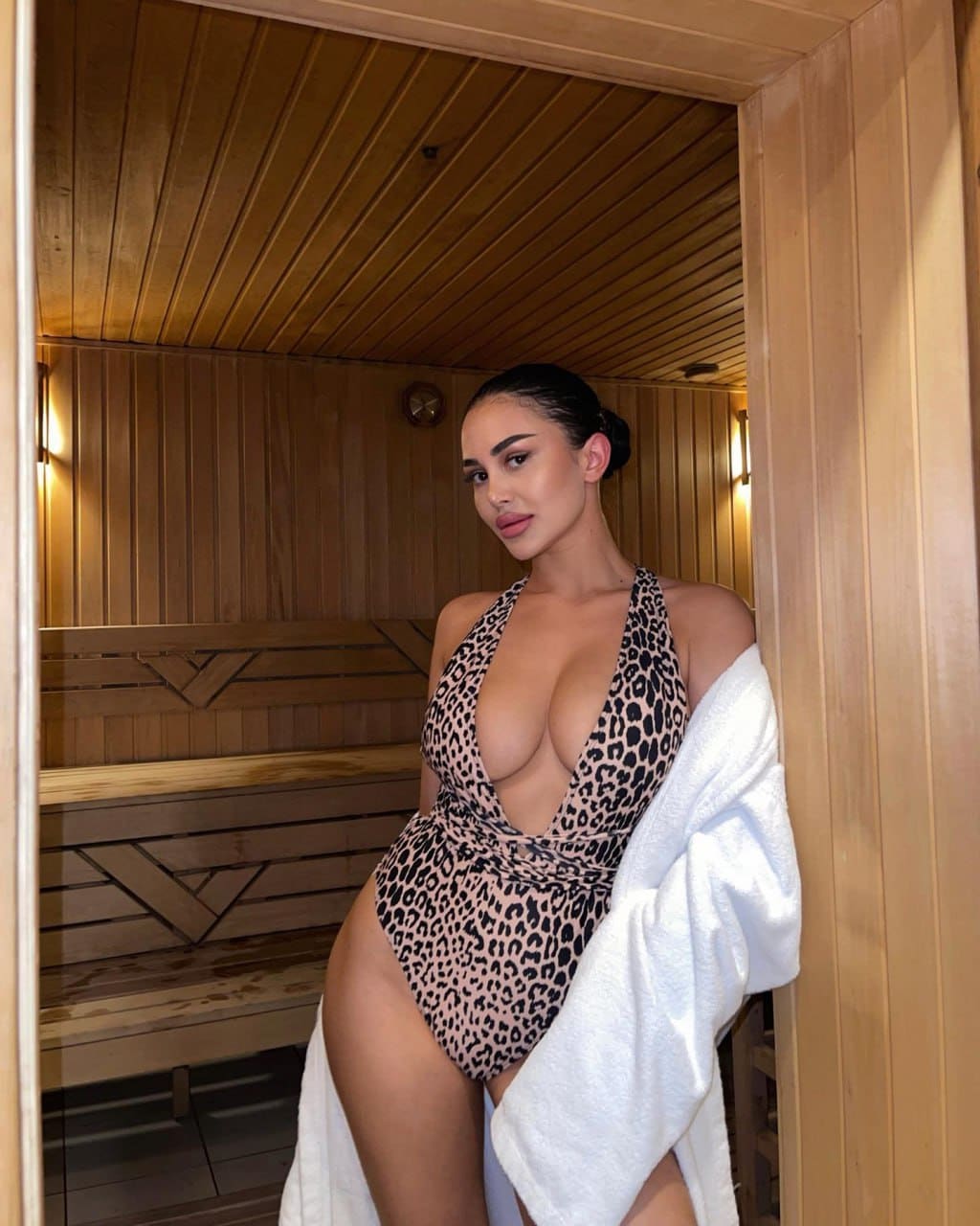A woman in a leopard-print halter swimsuit leans against a wooden sauna doorway, with a white towel over her shoulder and benches behind her.