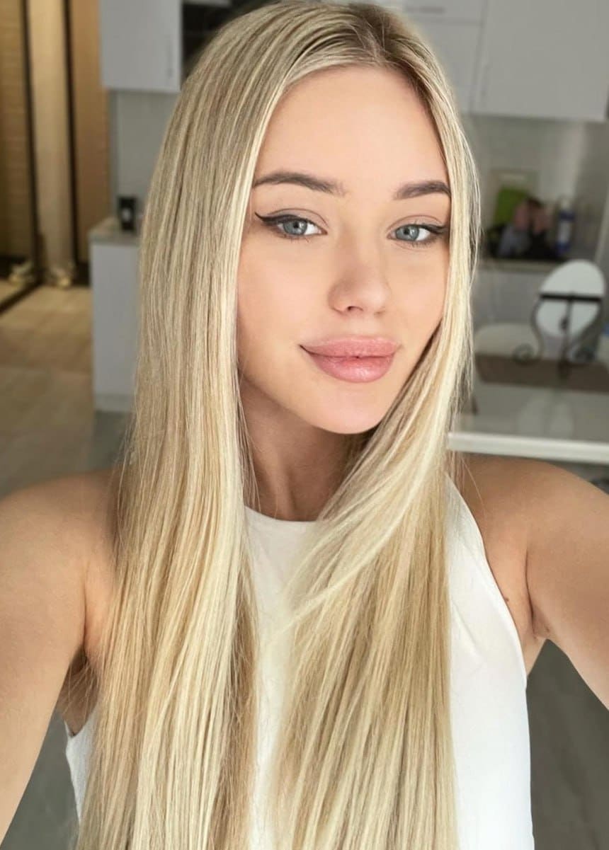 Luna with long blonde hair and winged eyeliner poses in a white sleeveless top in a bright room with white cabinets.