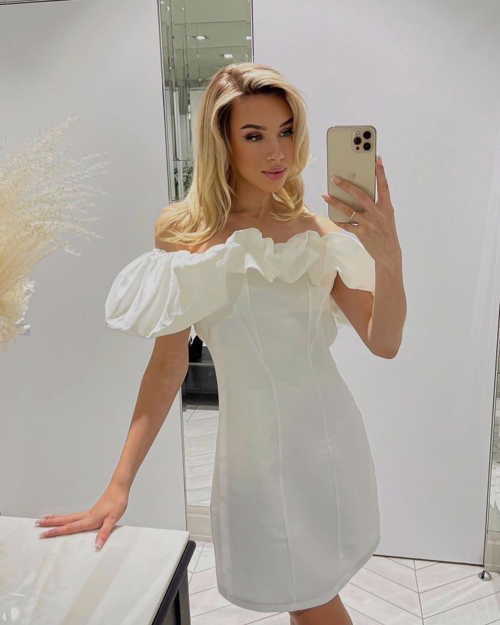 A mirror selfie features a woman in a white off-the-shoulder dress with ruffled neckline and puffed sleeves in a bright room.