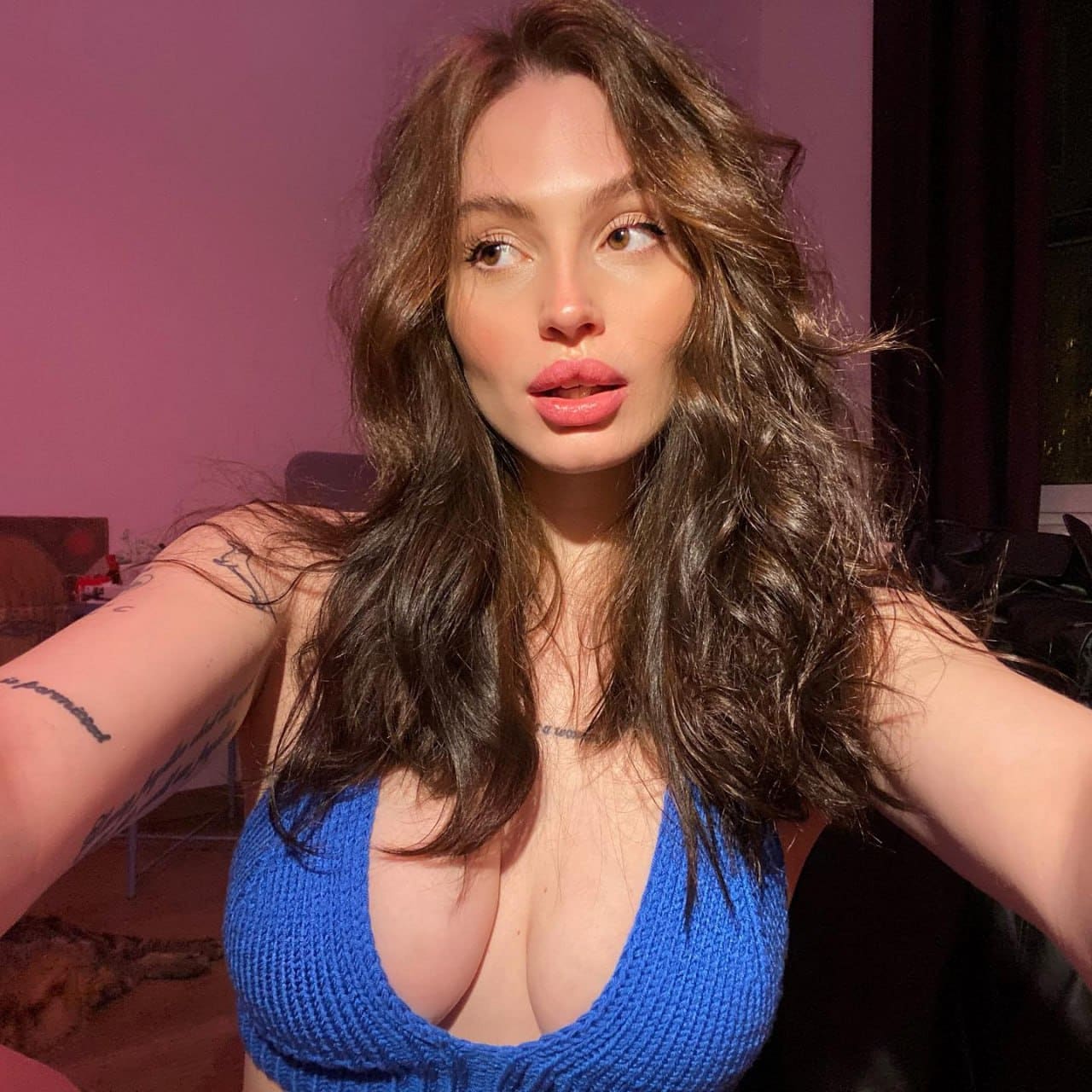 A woman takes a selfie in a blue ribbed crop top with a deep scoop neck, revealing arm and chest tattoos in a pink-lit room.