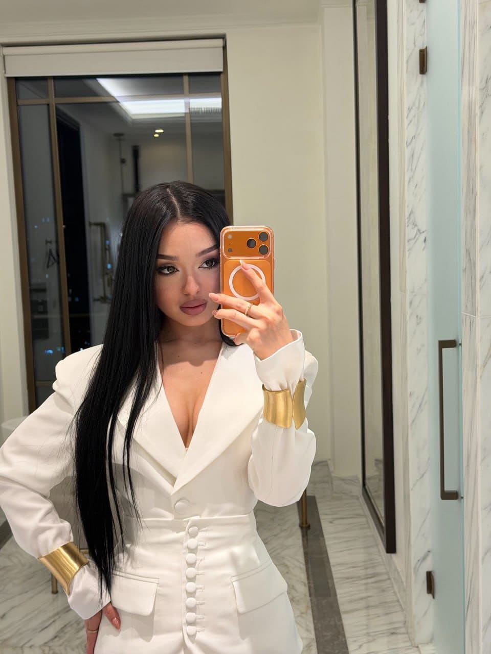 A mirror selfie shows a woman in a white blazer with a deep V-neck and gold bracelets, standing in a marble interior with a glass door.