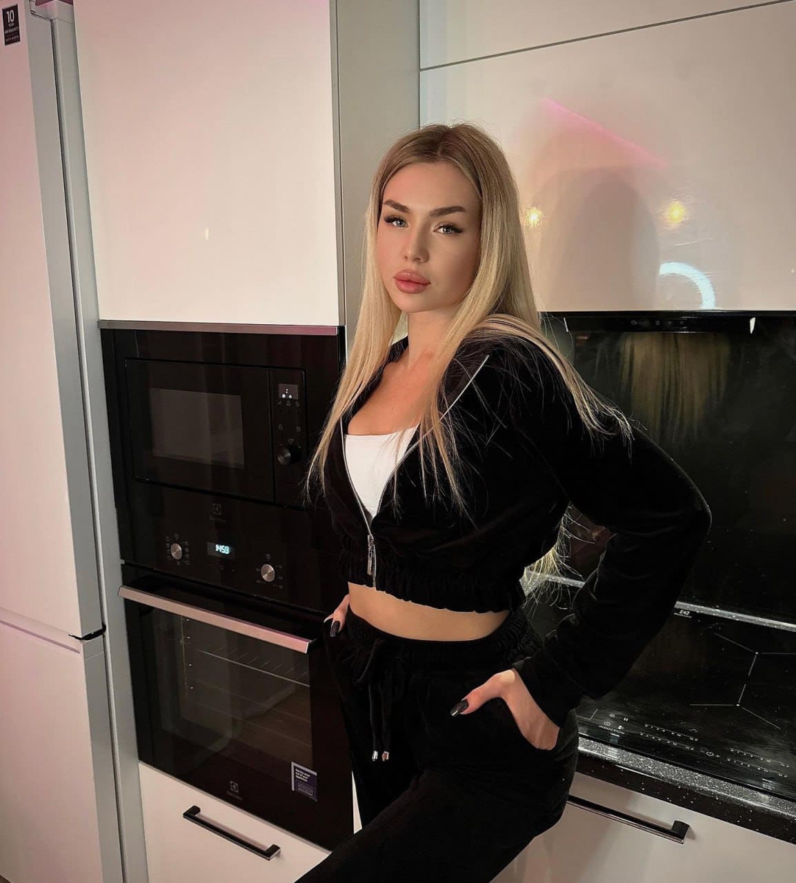 A blonde woman in a black cropped velvet jacket and pants stands in a modern kitchen with black appliances and white cabinets.