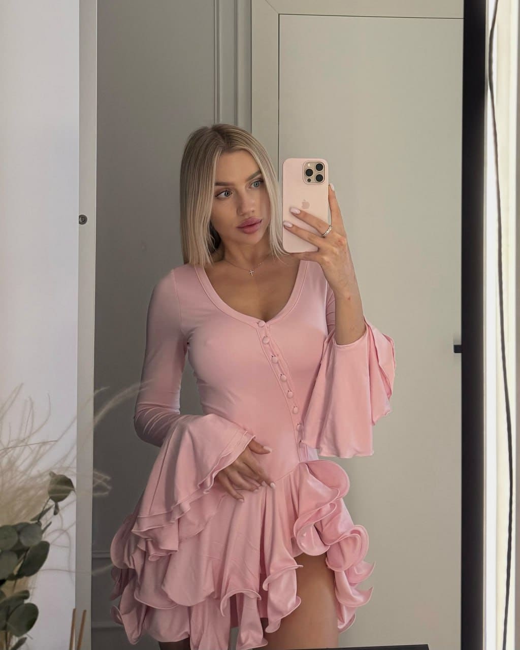 A blonde woman in a pink ruffled dress takes a mirror selfie indoors, with a white door and plant behind her.