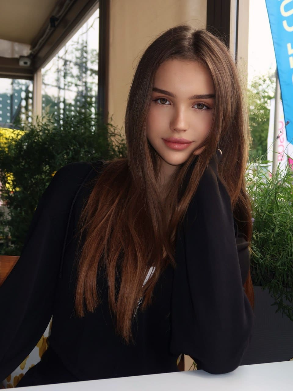 A woman sits at an outdoor white table wearing a black zippered top, with green plants and a window visible behind her.