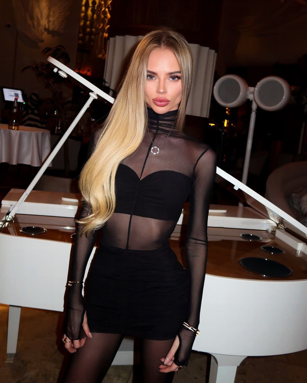 A woman in a sheer black top, bustier, and short skirt stands in an indoor venue near a white counter and lighting equipment.