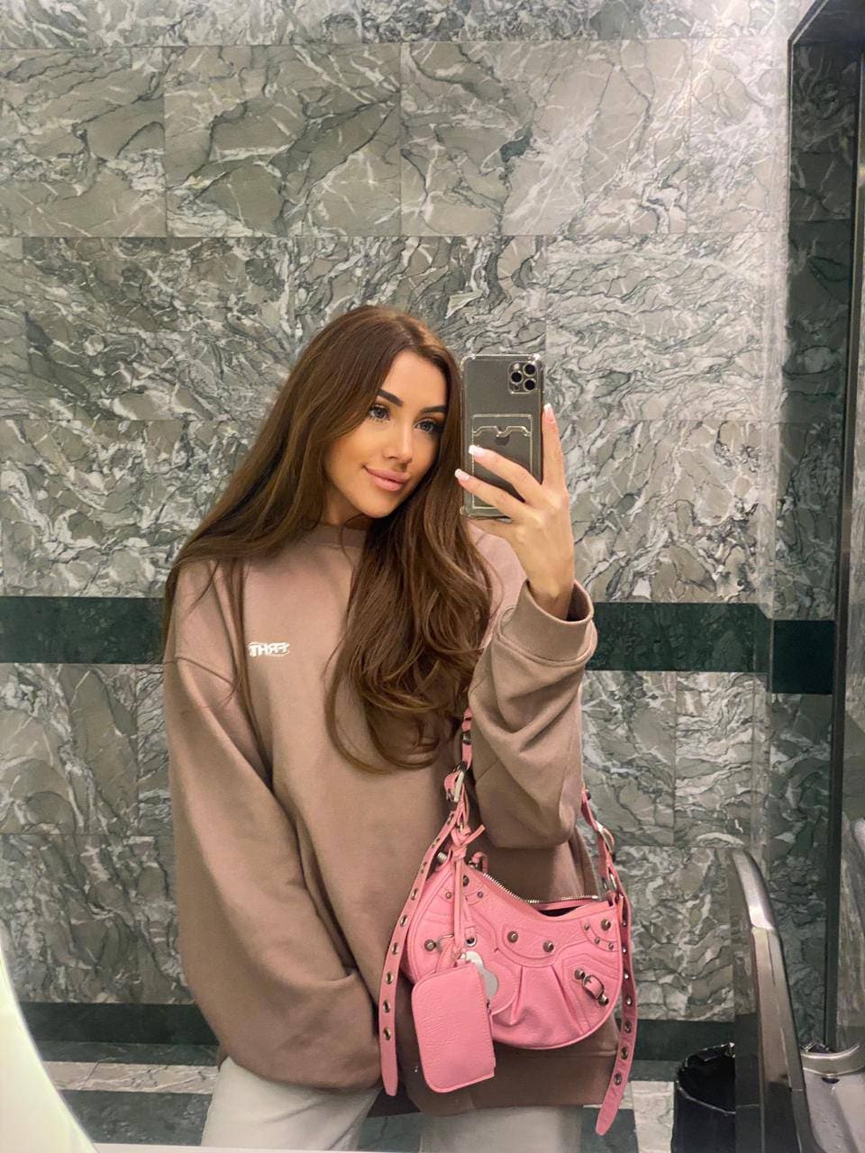 A mirror selfie shows a woman in a marble room wearing an oversized taupe sweatshirt and light pants, holding a phone and pink bag.