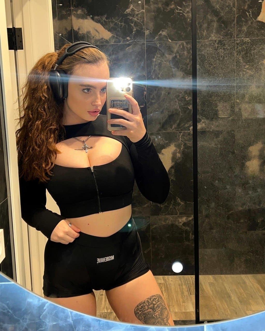 A mirror selfie shows a woman in headphones, a black zip crop top, and shorts, with a thigh tattoo visible against dark marble walls.