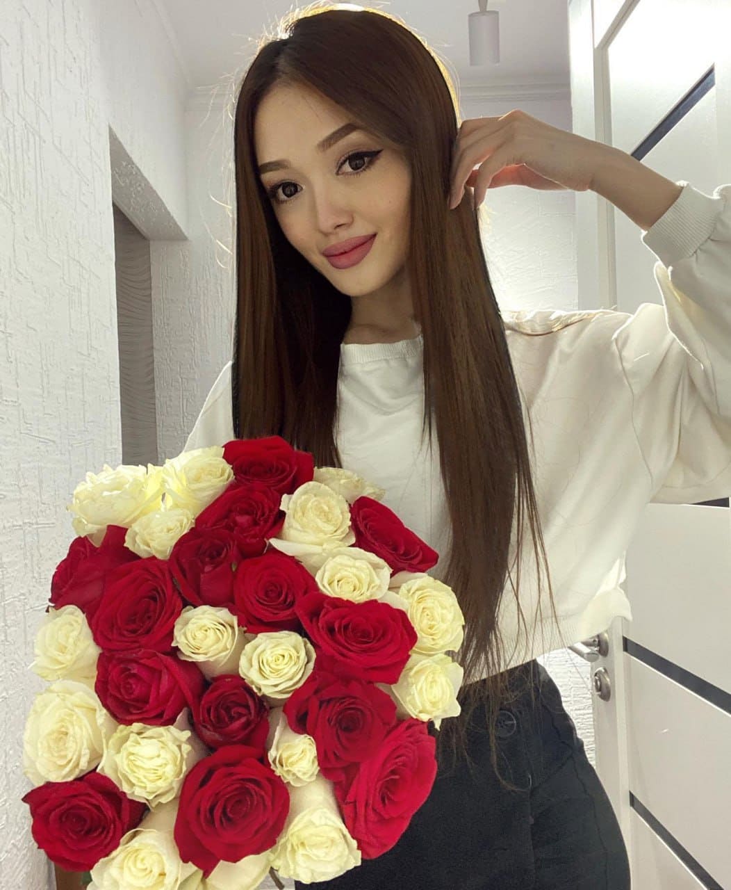 Vera stands indoors in a white long-sleeved top and dark pants, holding a large bouquet of red and white roses while touching her hair.