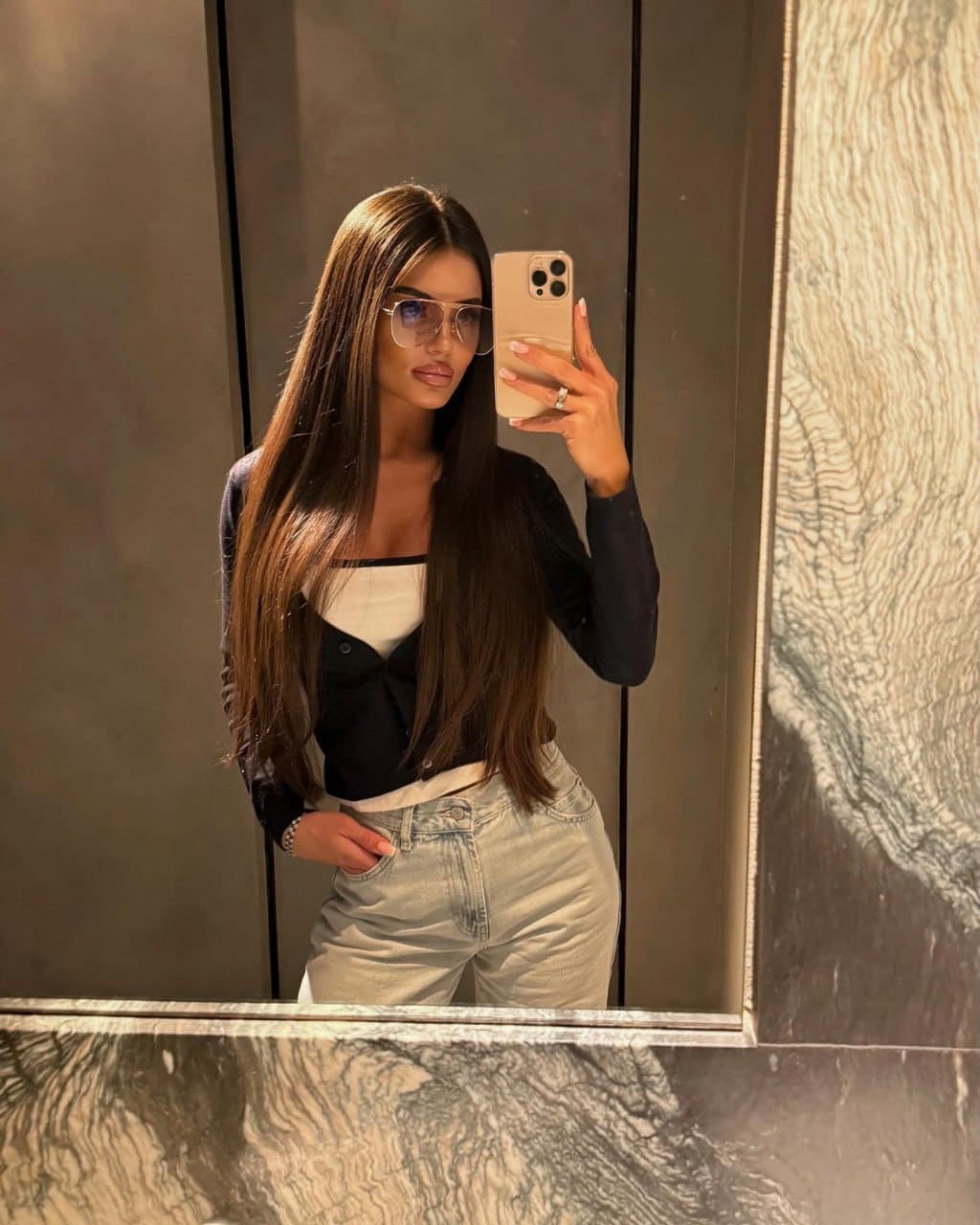 A woman in sunglasses, a black cardigan, and jeans takes a mirror selfie in a room with dark paneled walls.